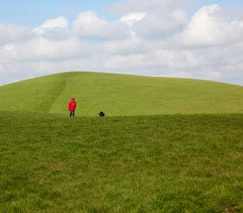 Lady Walking dog, green rolling hills This landscape photograph, taken around midday in early July, captures a scene at Snapes Point in Devon, England, United Kingdom. The image shows a person dressed in a red jacket walking a dog across the expansive green rolling hills, indicative of the summer season. The sky is partly cloudy, and the lush grass covers the gentle inclines of the countryside, providing a clear sense of the natural beauty found in this part of England. The photograph features people and animals as key elements, with the lady and her dog forming the main subjects against the rural backdrop typical of Devon.